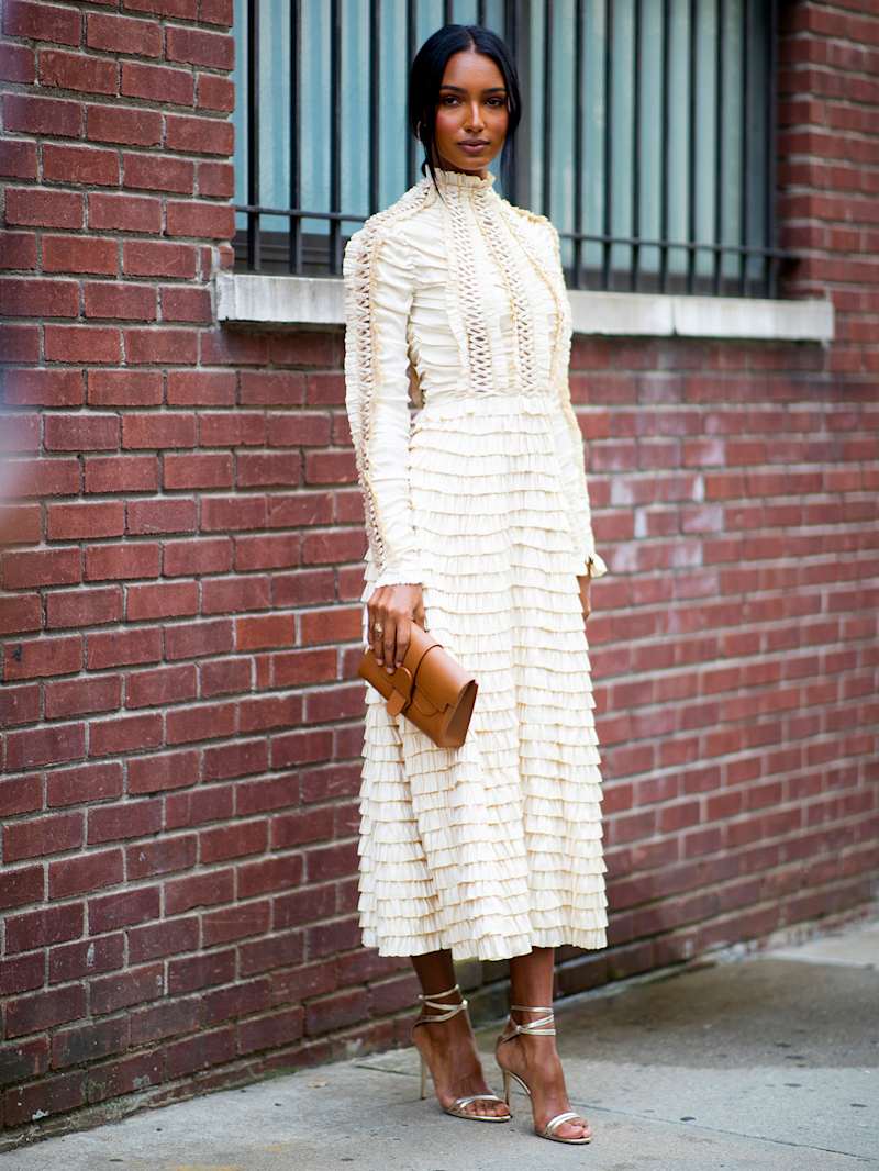 Frau trägt ein langes, hocheschlossenes Spitzenkleid in Creme und Metallic-Sandaletten, dazu eine cognacfarbe Clutch