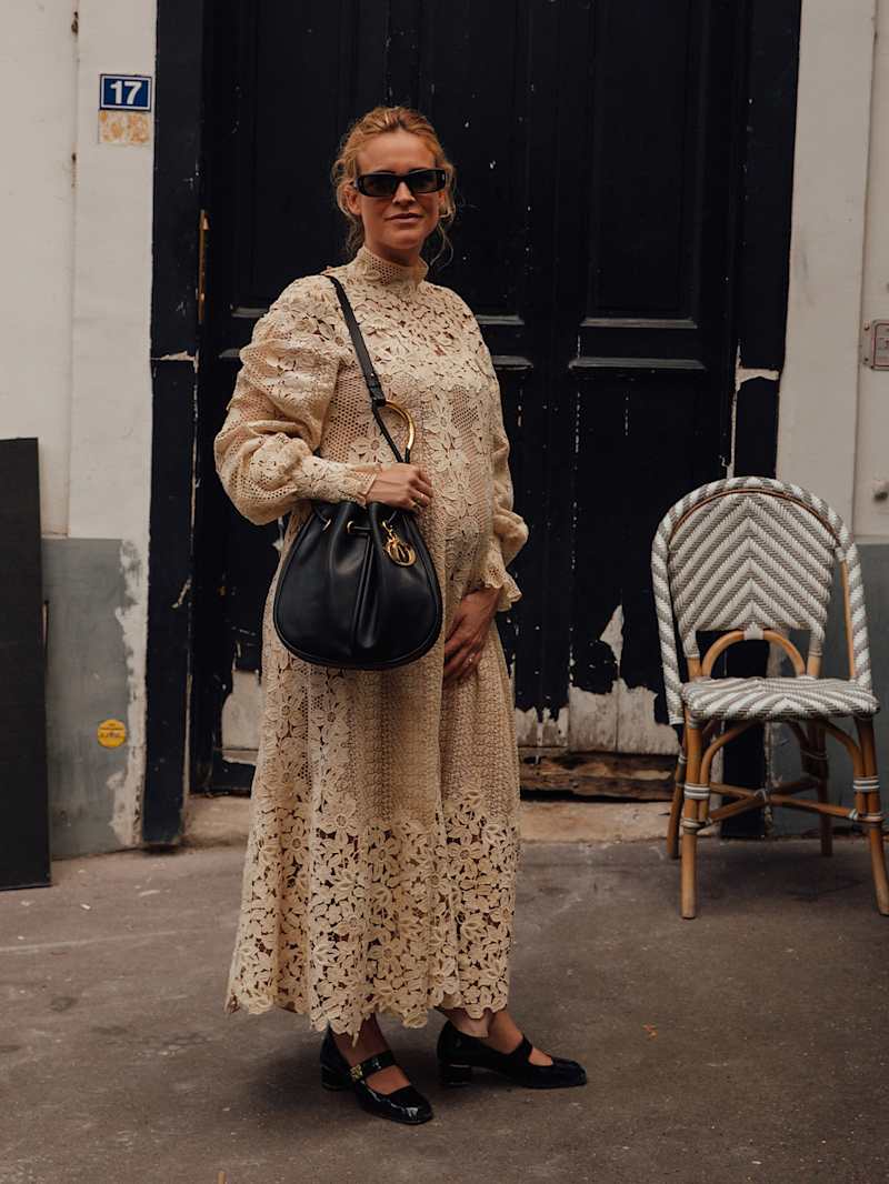 Frau trägt ein beiges Spitzenkleid, dazu schwarze, flache Mary-Janes und eine schwarze Hobo-Bag mit goldenen Applikationen