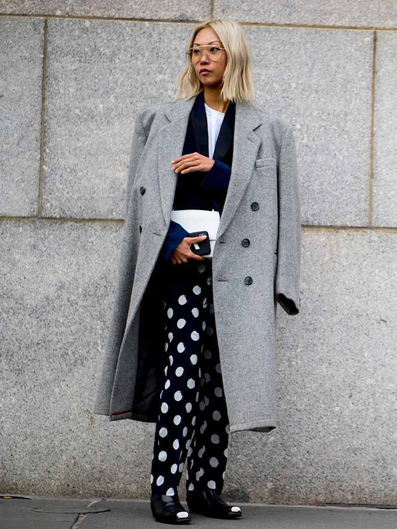 Frau trägt einen grauen Wollmantel zu einerm weißen Shirt und einer dunkelblauen Hose mit weißen Punkten
