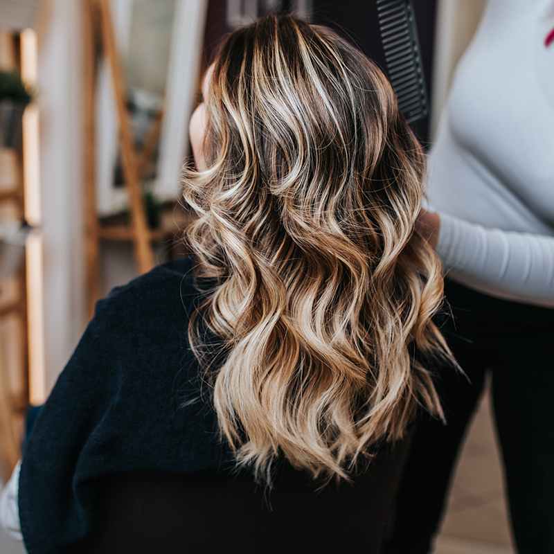 Frau mit blonder Balayage sitzt beim Friseur und lässt ihre Haare stylen