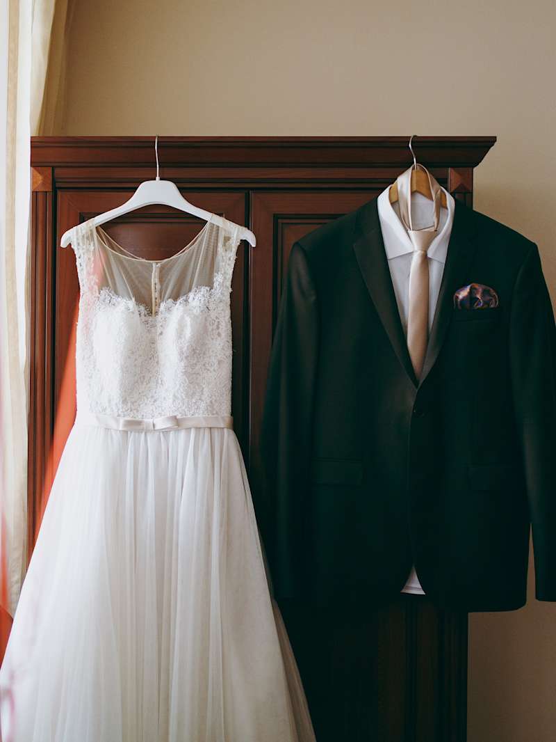 Ein Brautkleid und ein schwarzer Anzug hängen auf Kleiderbügeln an einem Schrank