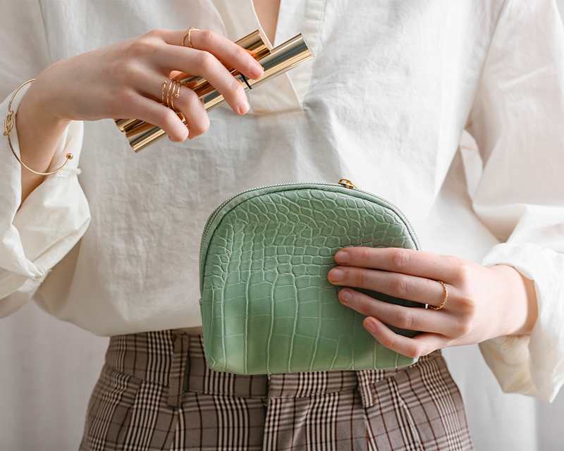 Eine Frau im weißen Hemd steckt ihre Make-up-Stifte in eine grüne Handtasche.
