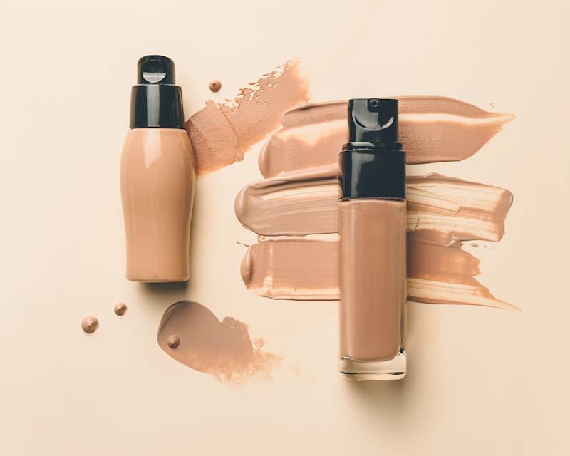 Aus drei aufeinander gestellten Glasflaschen läuft flüssiges Make-up auf den Stoff.