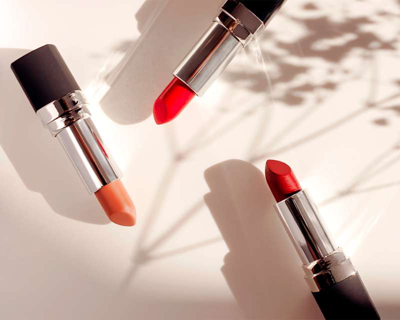 Zwei rote und ein orangefarbener geöffneter Lippenstift, die auf einer hellbeigen Unterlage Schatten werfen.