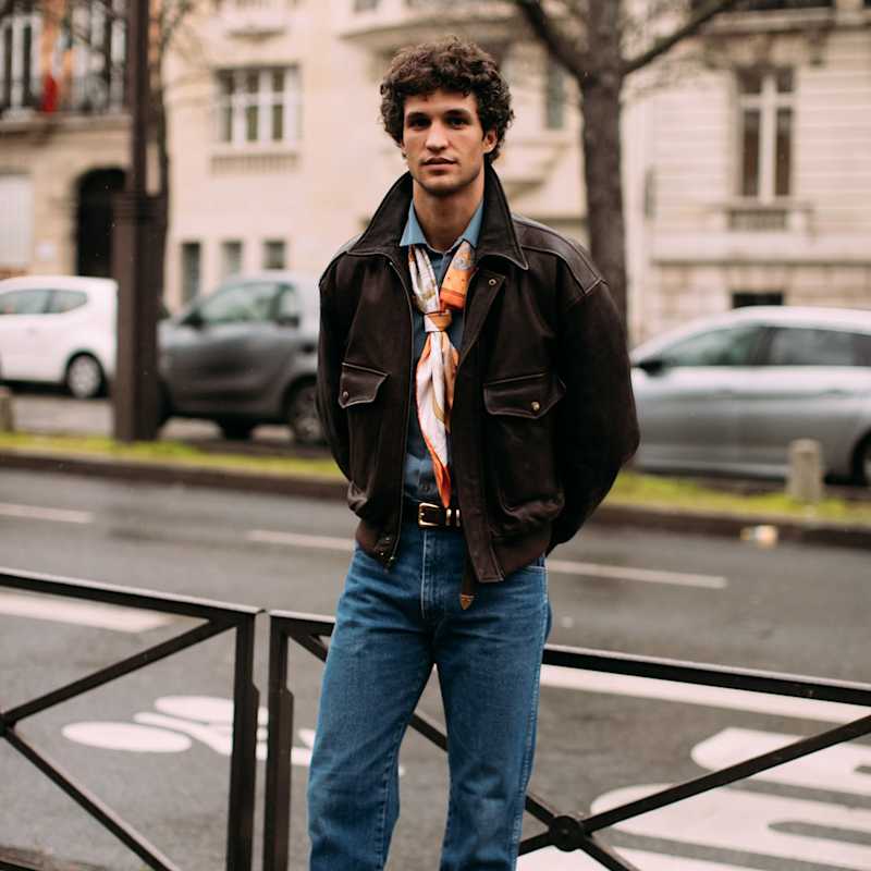 Mann trägt eine Lederjacke zu Jeans und bunten Tuch und steht an einer Straße