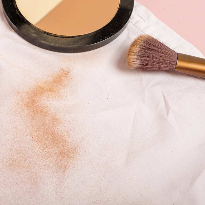 Weißes Kleidungsstück mit Puderflecken neben Make-up-Pinsel und Puderdose.