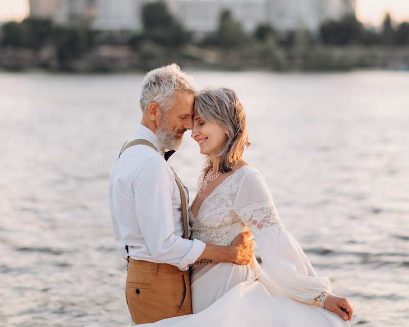 Frau trägt ein weißes Kleid und der Mann, der sie an der Taille festhält, trägt eine beige Hose, ein weißes Hemd und braune Hosenträger
