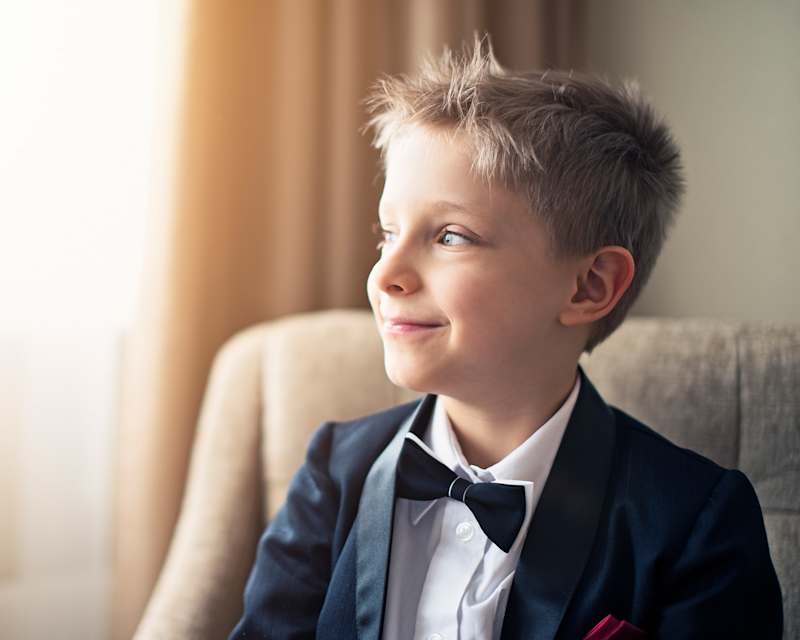 Junge in einem dunkelblauen Sakko und einer Fliege sitzt auf einem Sessel