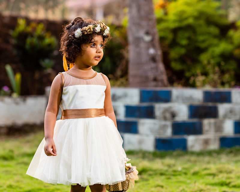 Kleines Mädchen in einem weißen Kleid trägt einen Blumenhaarkranz und hält einen Korb in der Hand