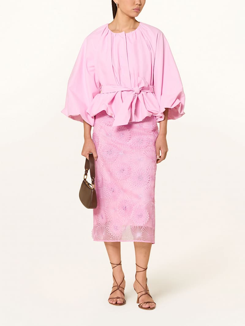 Frau trägt rosafarbene Bluse zu floralem Spitzen-Bleistiftrock mit Riemchensandalen und Handtasche