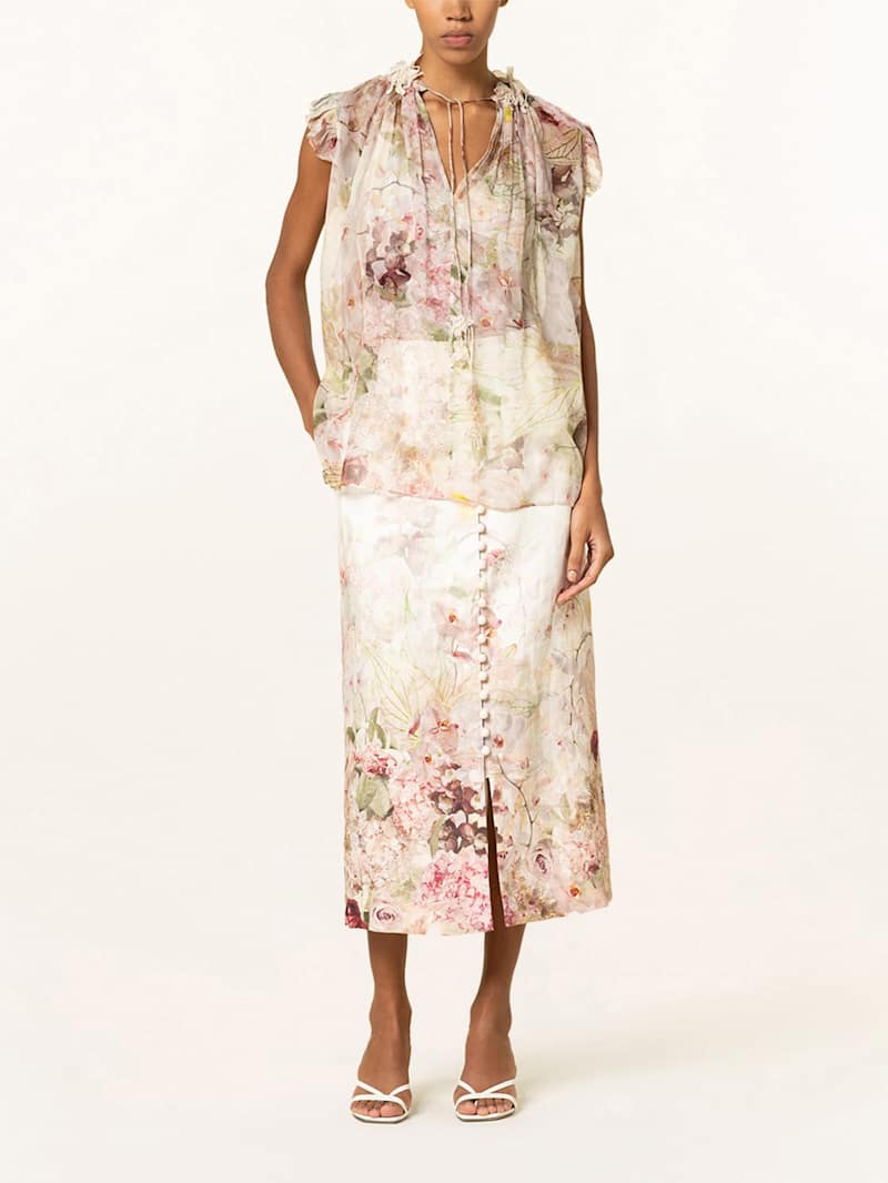 Frau trägt florale, semitransparente Bluse mit Rüschen-Details zu passendem, knielangem Rock mit Knopfleiste.