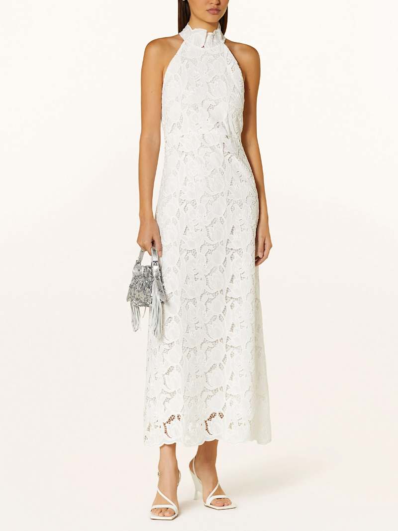 Frau trägt ein knöchellanges weißes Spitzenkleid mit Neckholder und einer silbernen Handtasche