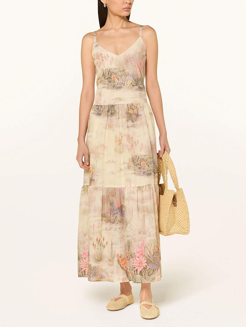 Frau trägt ein fließendes, knöchellanges Kleid mit floralen Muster, dazu eine Basttasche und Ballerinas