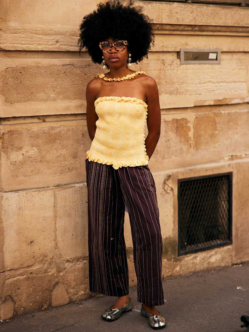 Frau trägt ein gelbes trägerloses Top zu einer weiten Hose und silbernen Tabi-Shoes, dazu Sonnenbrille und Ohrringe mit großen Perlen