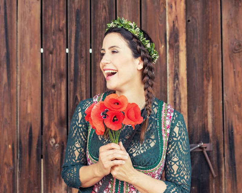 Frau in einem grünen Dirndl mit einer geflochtenen Dirndl-Frisur hält einen Mohnblumenstrauß in den Händen 