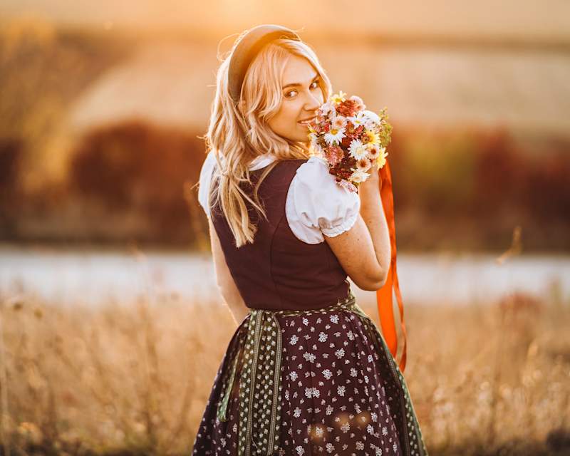 Frau mit langen blonden Haaren steht auf einem Feld, trägt ein Dirndl und hält einen Blumenstrauß 