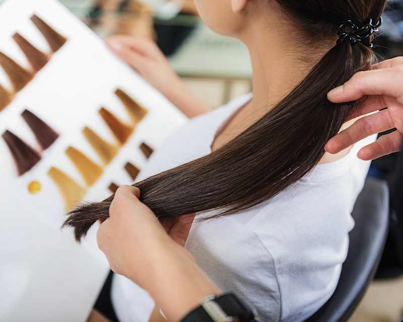 Frau sitzt beim Friseur mit dem Rücken zur Kamera und betrachtet eine aufgeklappte Farbpalette mit verschiedenen Haarsträhnen