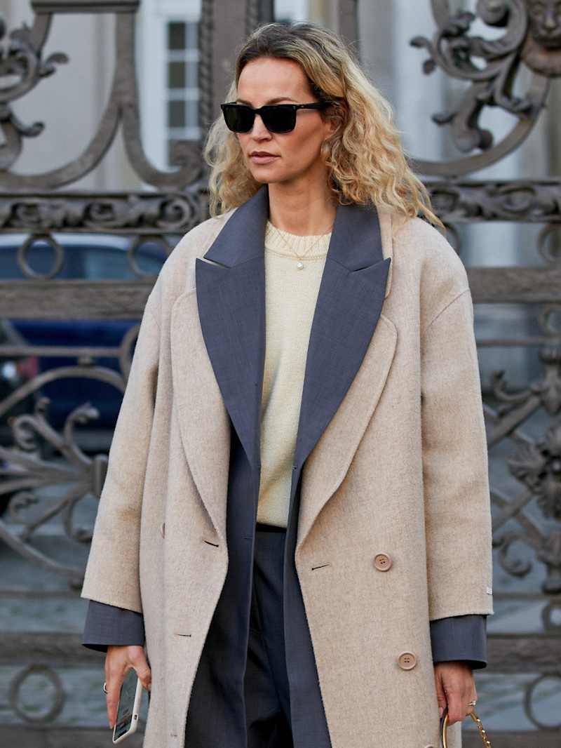 Eine Frau mit blonden Locken trägt einen beigen Wollmantel über einem grauen Blazer und einem gelben Shirt