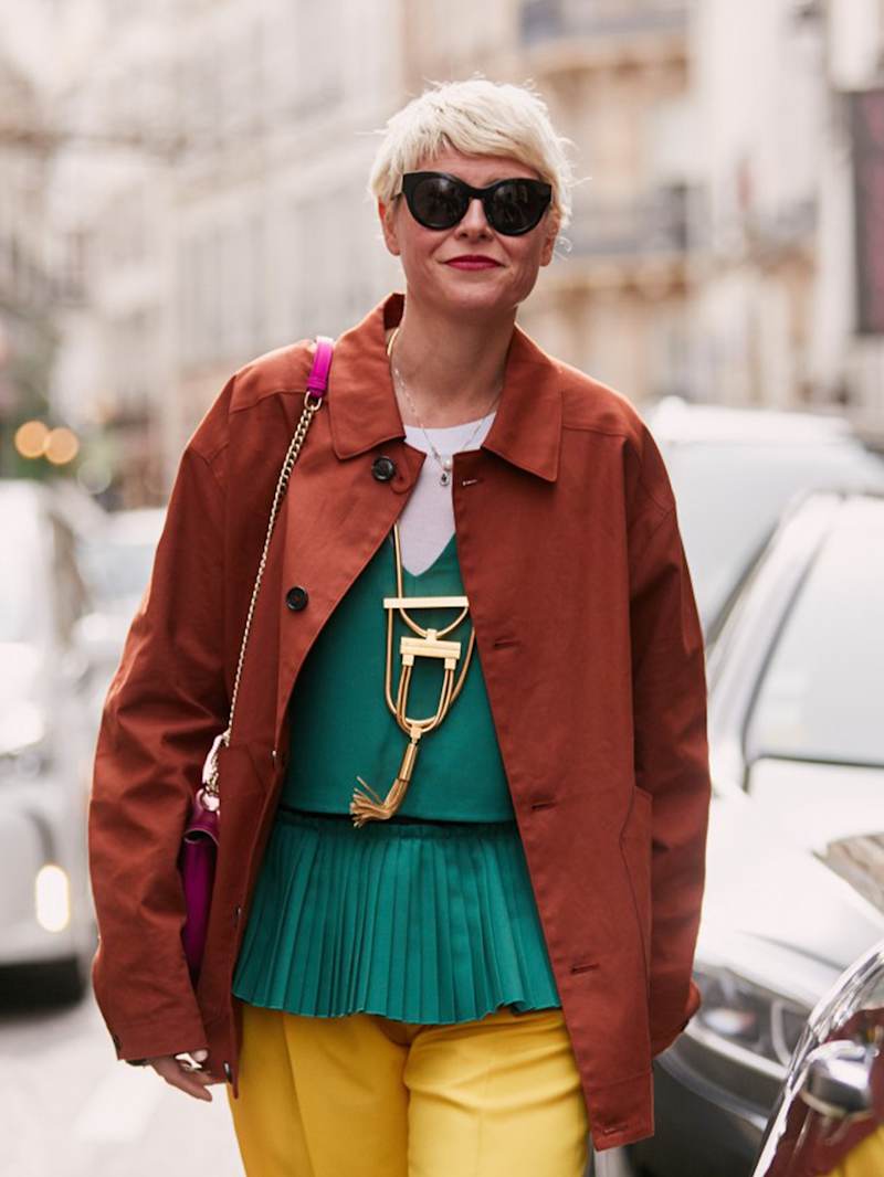 Frau mit der roten Jacke und gelber Hose trägt ihre blonden Haare als modernen Pixie-Cut.