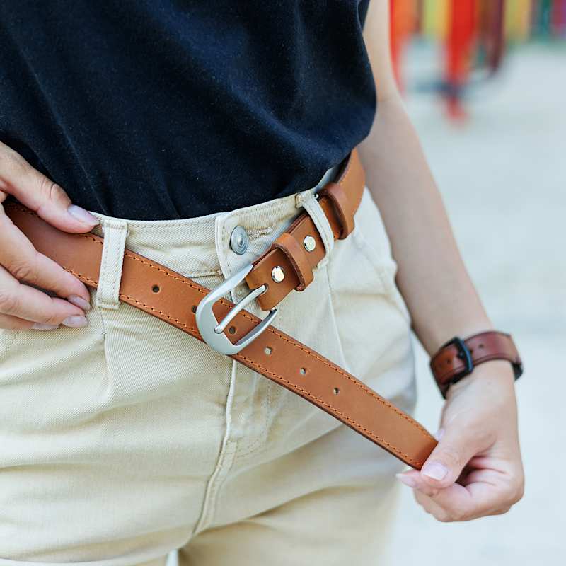 Nahaufnahme einer Frau, die einen braunen Ledergürtel über einer beigefarbenen High-Waist-Hose schließt