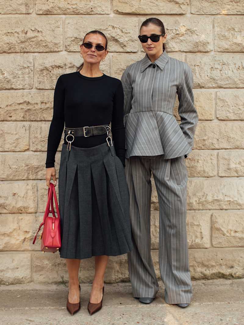 Zwei Frauen mit Sonnenbrillen präsentieren Herbst-Outfits vor einer Natursteinwand: links ein schwarzes Langarmoberteil mit grauem Faltenrock und roter Handtasche, rechts ein grauer Nadelstreifen-Hosenanzug.