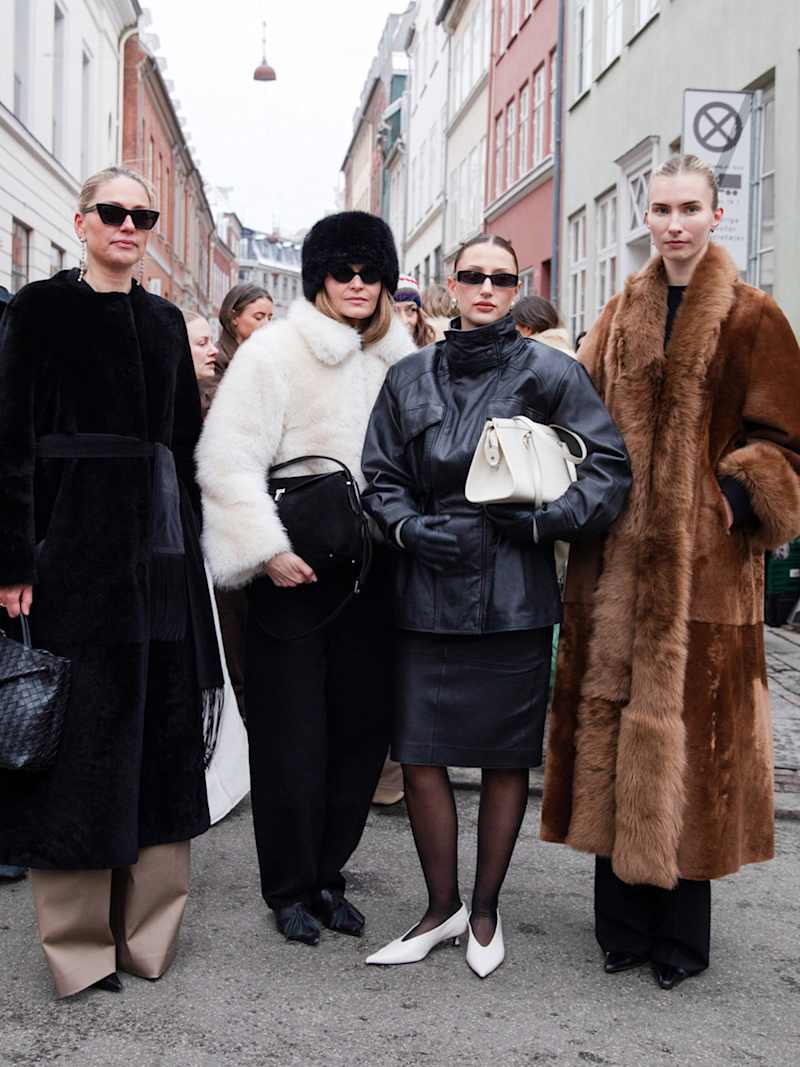 Vier Frauen in eleganten Winteroutfits stehen auf einer Straße und tragen unter anderem schwarze Mäntel, eine schwarze Lederjacke und einen langen braunen Mantel.