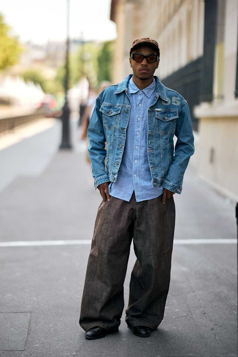 Mann steht auf einer Straße und trägt ein Jeansjacke zu einer grauen Jeanshose, dazu ein Cap und Sonnenbrille. 