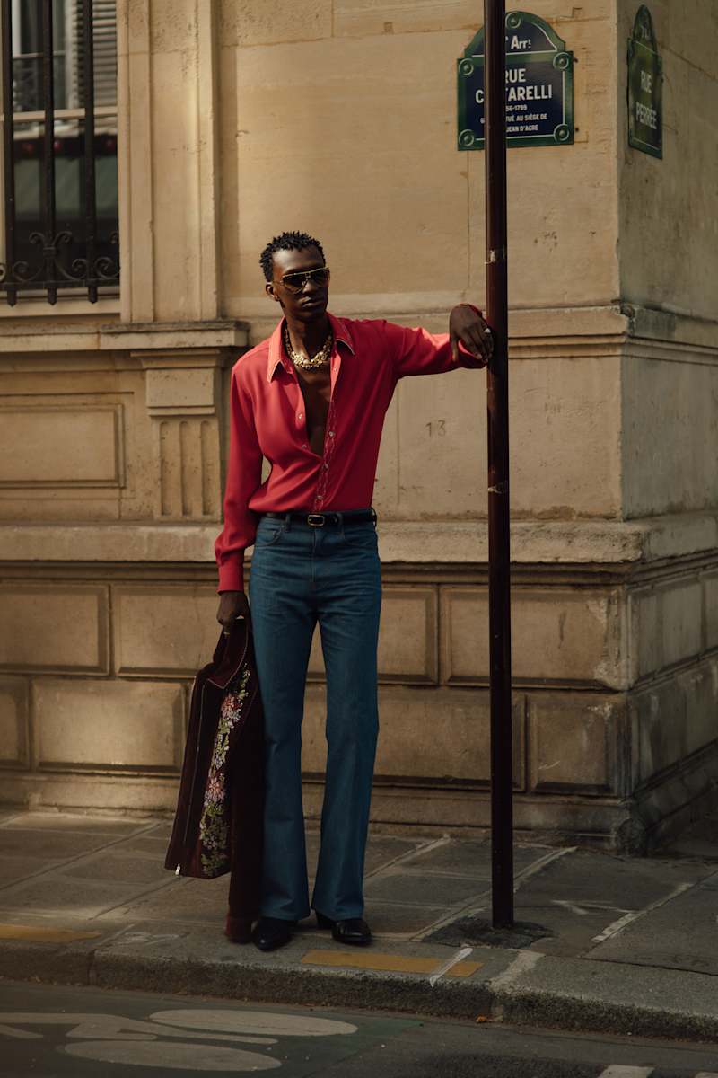 Mann trägt ein rotes Hemd zu einer blauen Skinny Jeans, in der Hand hält er eine braune Jacke.