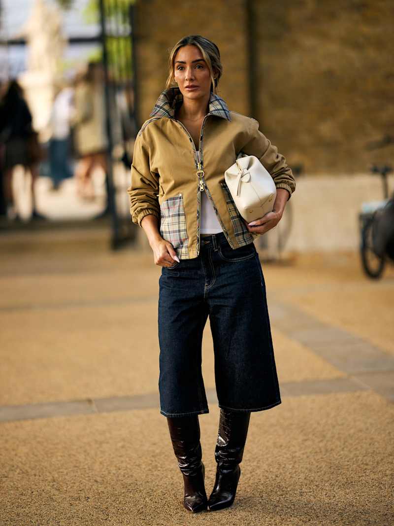 Frau trägt eine beige Jacke zu einer kurzen Culotte aus Indigo Denim, dazu eine cremefarbene Tasche unter dem Arm und Stiefel. 