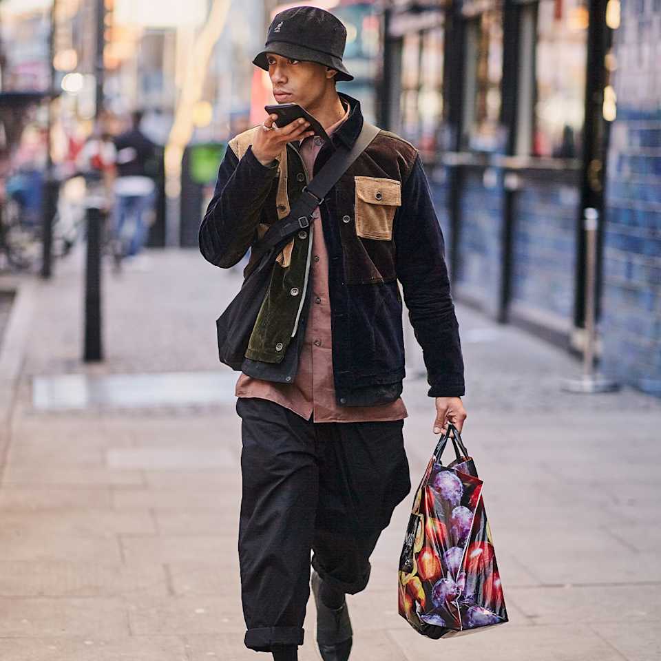 Mann trägt eine schwarze Jacke, eine lockere Stoffhose und einen Bucket Hat im Modern Utility Style.