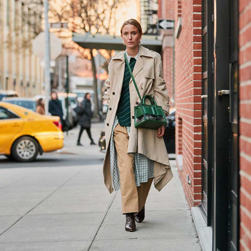 Frau trägt in einen beigen Trenchcoat über einem karierten Hemd und grünem Strickpullover als Teil des Layering-Looks