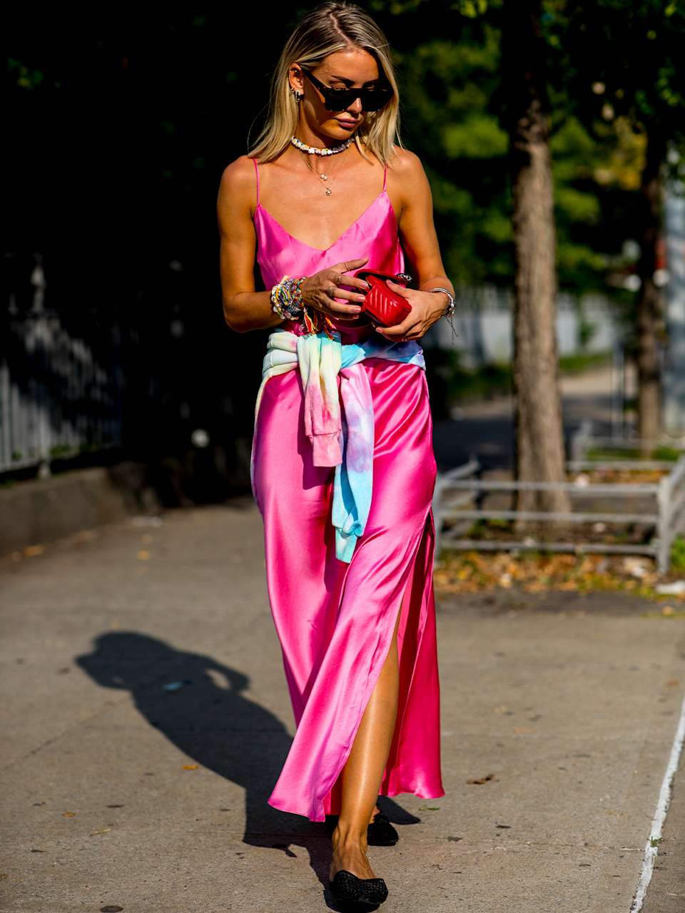 Frau in rosarotem Slip-Dress kontrolliert im Gehen ihre rote Handtasche mit Blick durch Sonnenbrille.