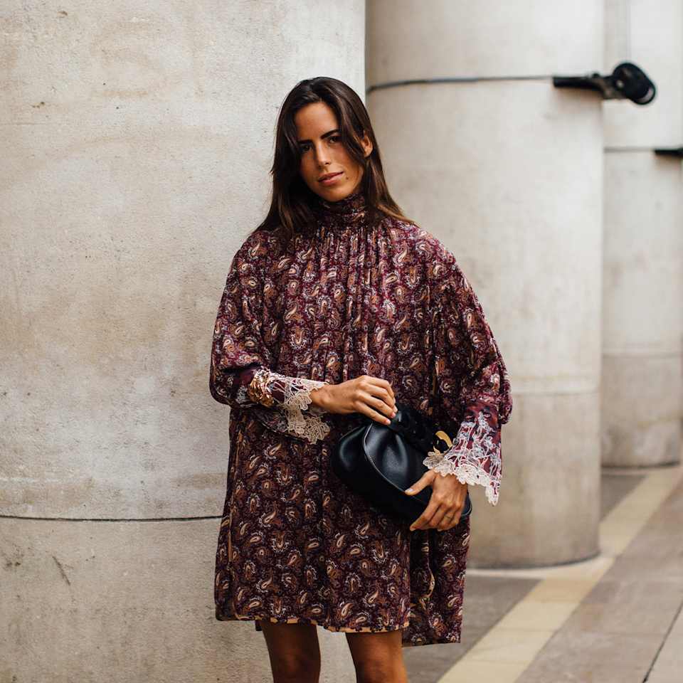 Frau in einem hochgeschnittenen Kleid mit Paisleymuster in der Farbe Bordeauxrot hält eine schwarze Leder-Clutch in der Hand