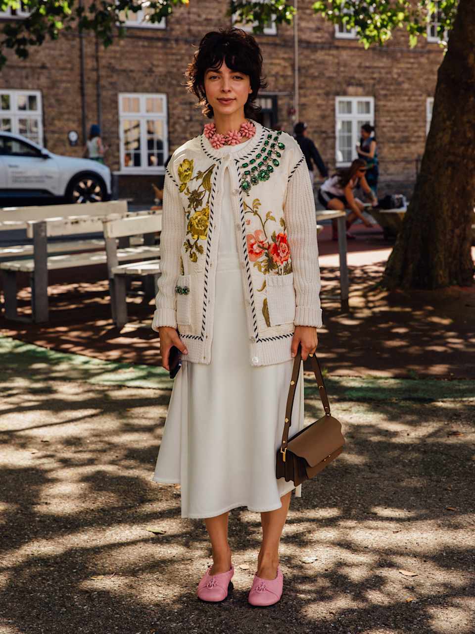 Frau trägt einen weißen Cardigan mit floralen Muster über einem weißen Kleid zu rosafarbenen Mules und einer braunen Handtasche