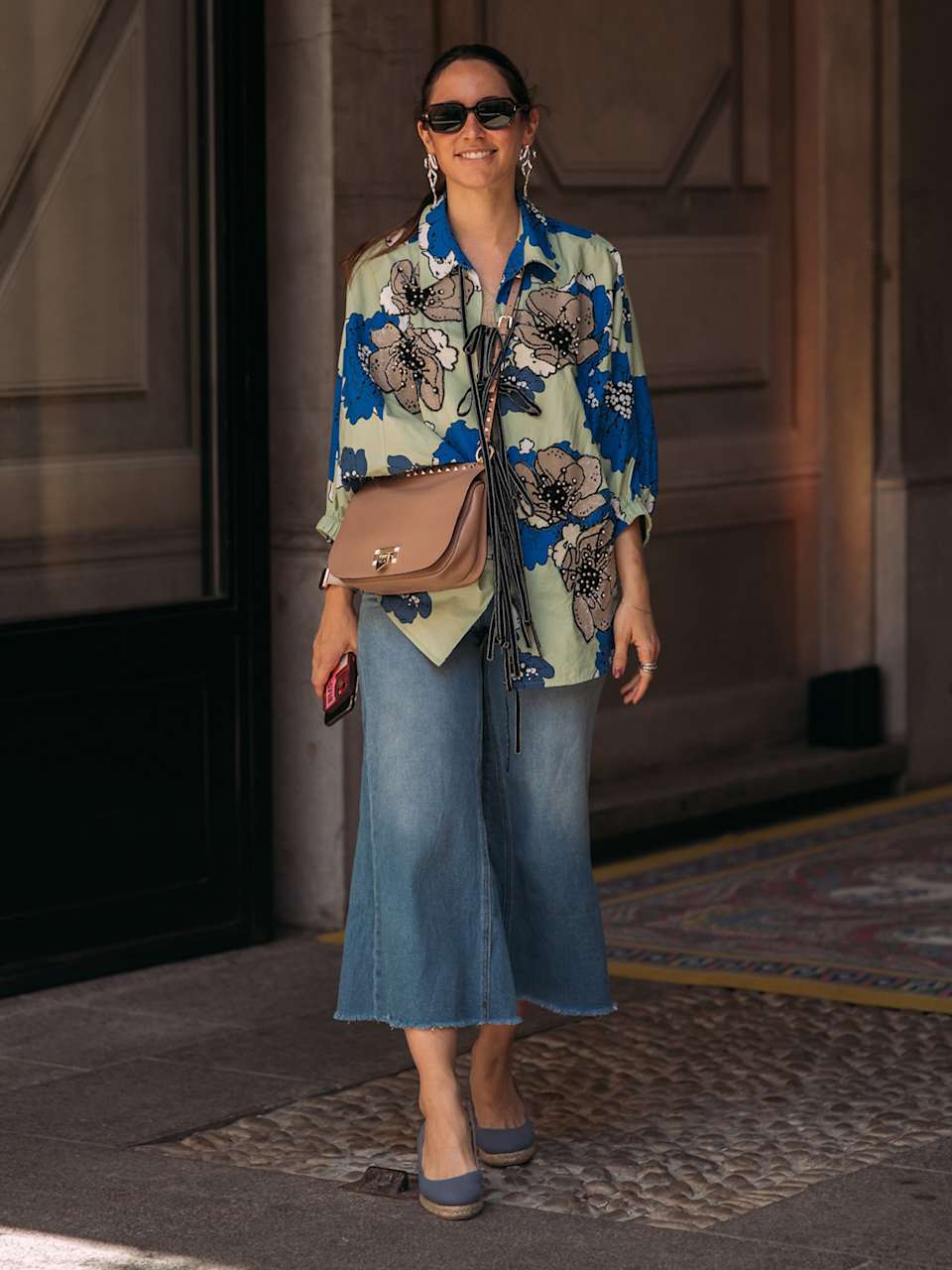 Frau trägt eine Hemdbluse mit Blumenmuster zu einer Jeans-Culotte und blauen Wedges, dazu eine braune Umhängetasche