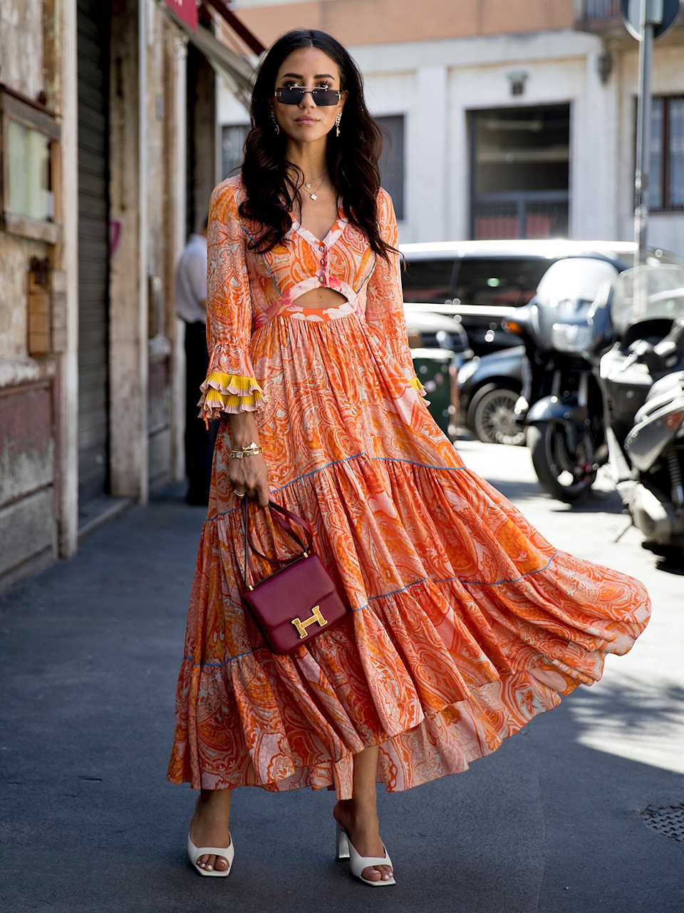 Frau trägt ein Maxi-Kleid in der Farbe Orange mit Paisleymuster, dazu weiße Mules mit Absatz und eine dunkelrote Umhängetasche