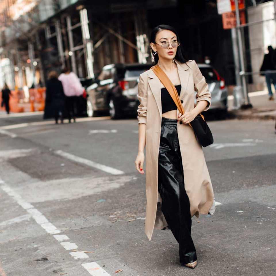 Frau trägt zu einer schwarzen Lederhose ein Tube-Top sowie Helle Pumps und einen beigen Trenchcoat