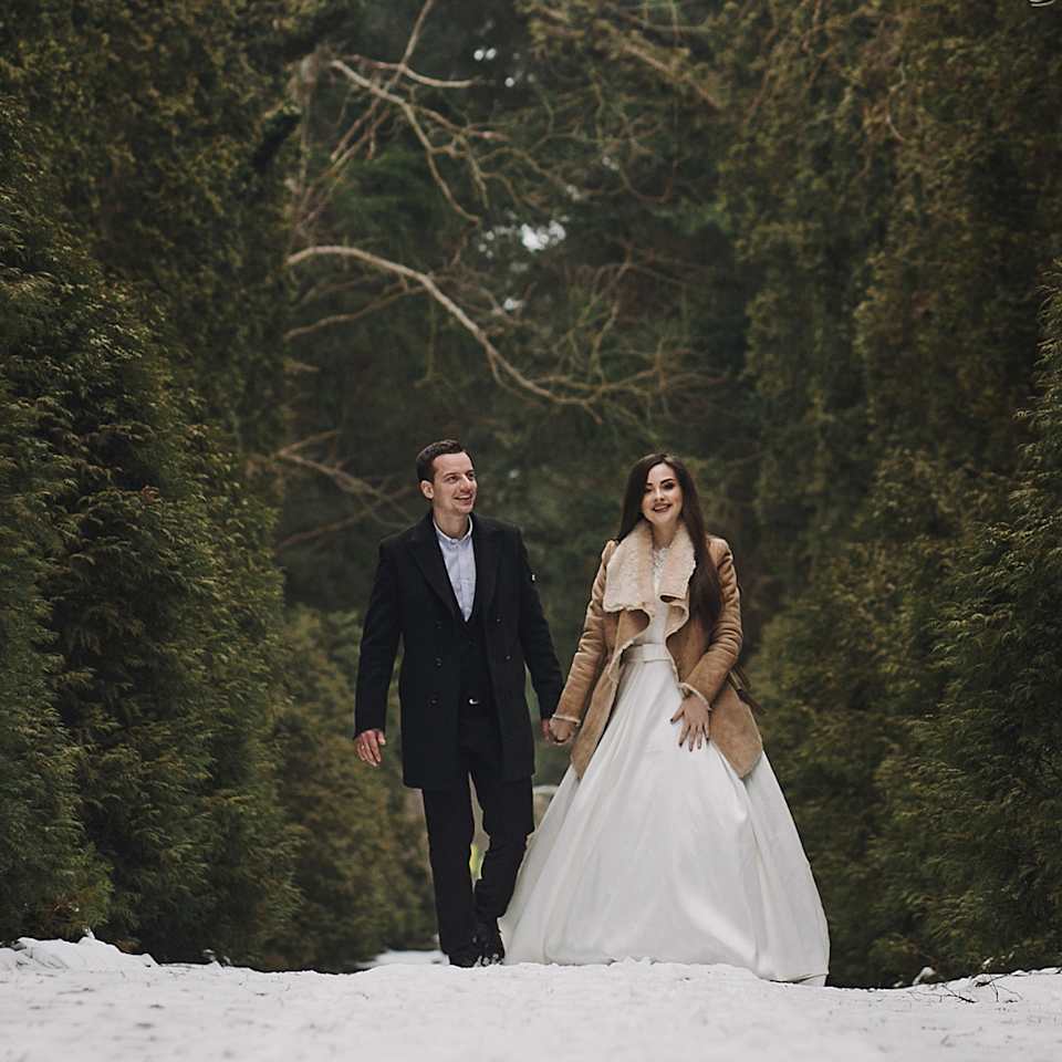 Brautpaar steht im Wald und es liegt Schnee, sie tragen winterliche Hochzeits-Outfits und Mäntel