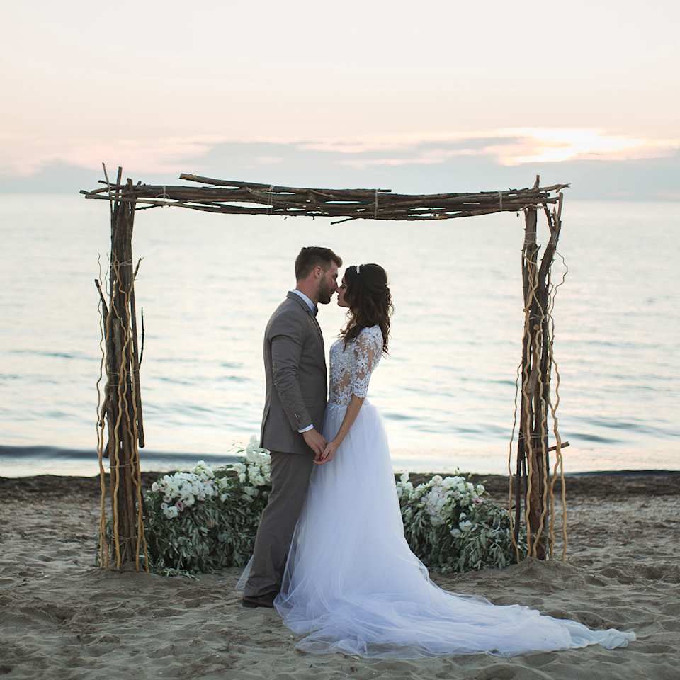 Brautpaar steht am Strand und hält sich an den Händen, im Hintergrund sieht man das Wasser