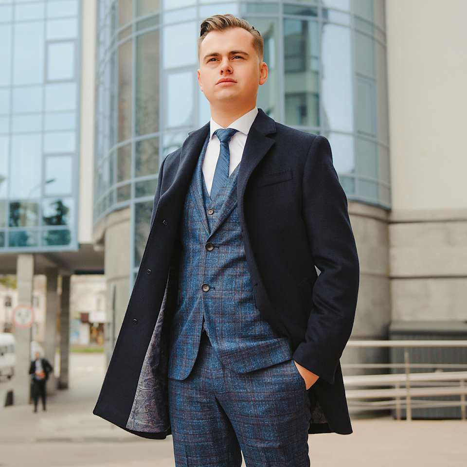 Herr in blau kariertem Anzug mit Weste, Krawatte und dunklem Mantel vor modernem Gebäude