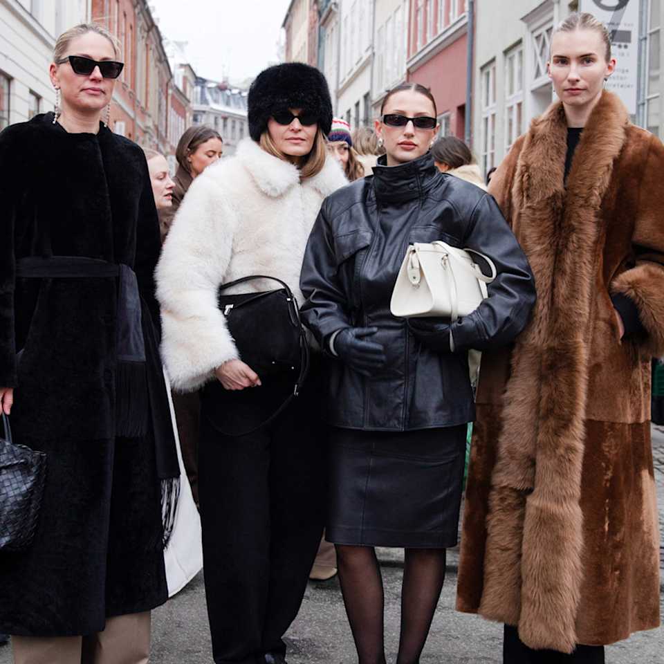 Vier Frauen in eleganten Winteroutfits stehen auf einer Straße und tragen unter anderem schwarze Mäntel, eine schwarze Lederjacke und einen langen braunen Mantel.