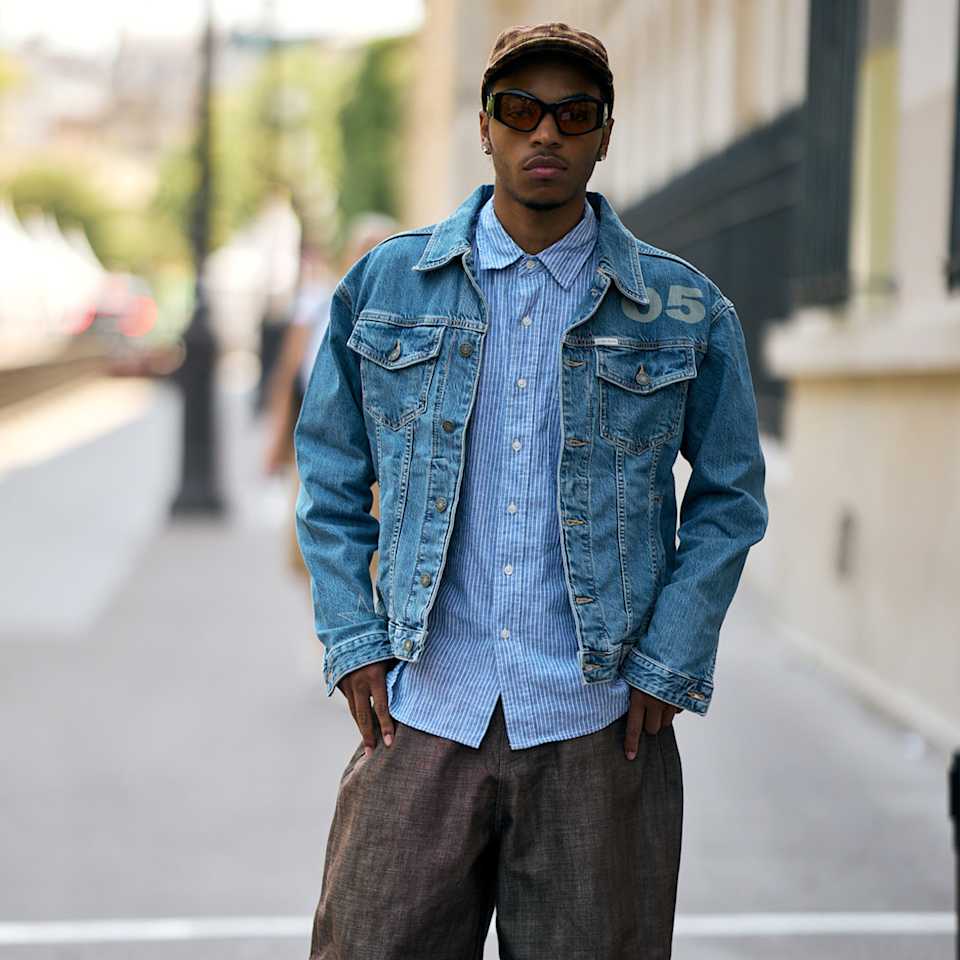 Mann steht auf einer Straße und trägt ein Jeansjacke zu einer grauen Jeanshose, dazu ein Cap und Sonnenbrille. 