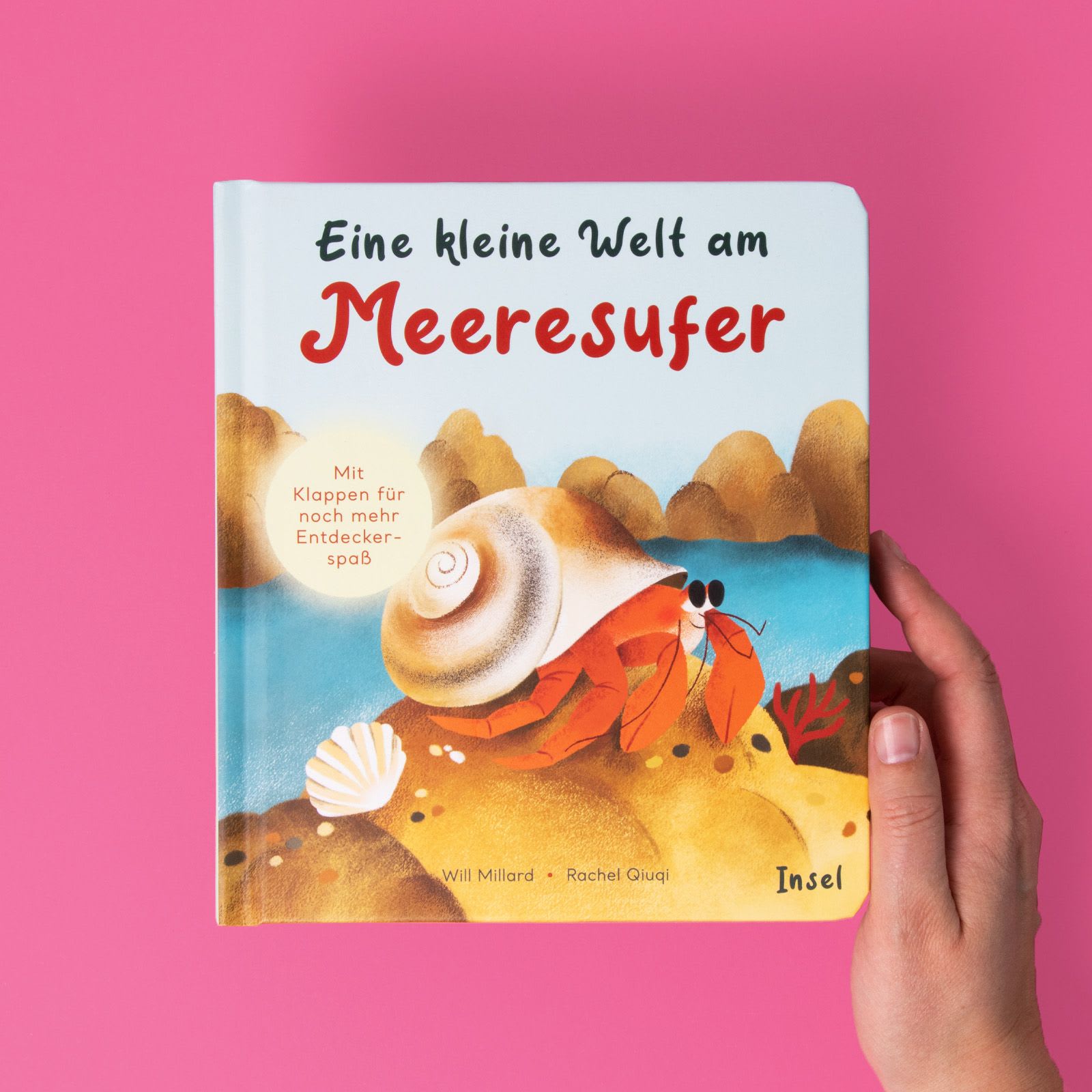 Eine Hand hält das Kinderbuch »Eine kleine Welt am Meeresufer« von Will Millard vor einem pinken Hintergrund.