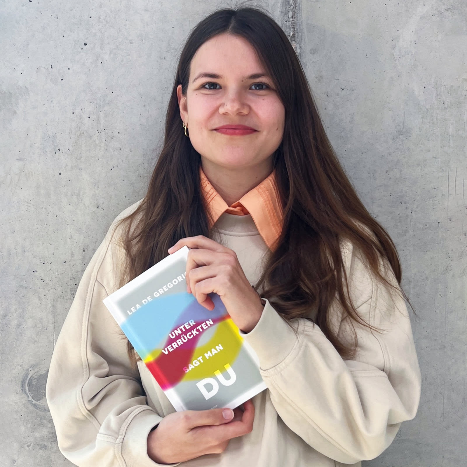 Die Autorin Lea De Gregorio steht lächelnd vor einer grauen Betonwand und hält ihr Buch »Unter Verrückten sagt man du« in den Händen.
