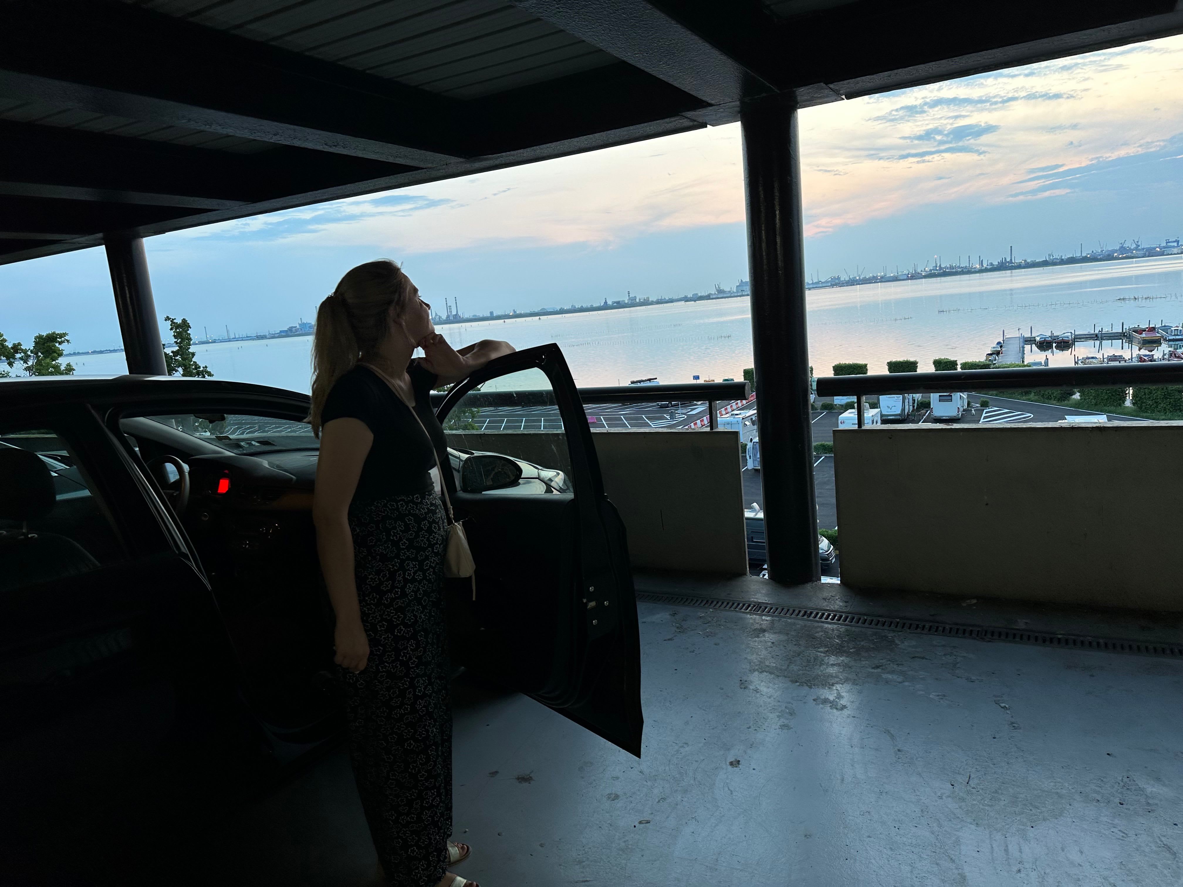 Eine Frau steht an der offenen Beifahrertür eines Autos, das sich auf einem Parkdeck eines Schiffs befindet, und blickt in Richtung des umgrenzenden Meers. Am Horizont ist Venedig zu sehen. Copyright: Yannic Han Biao Federer
