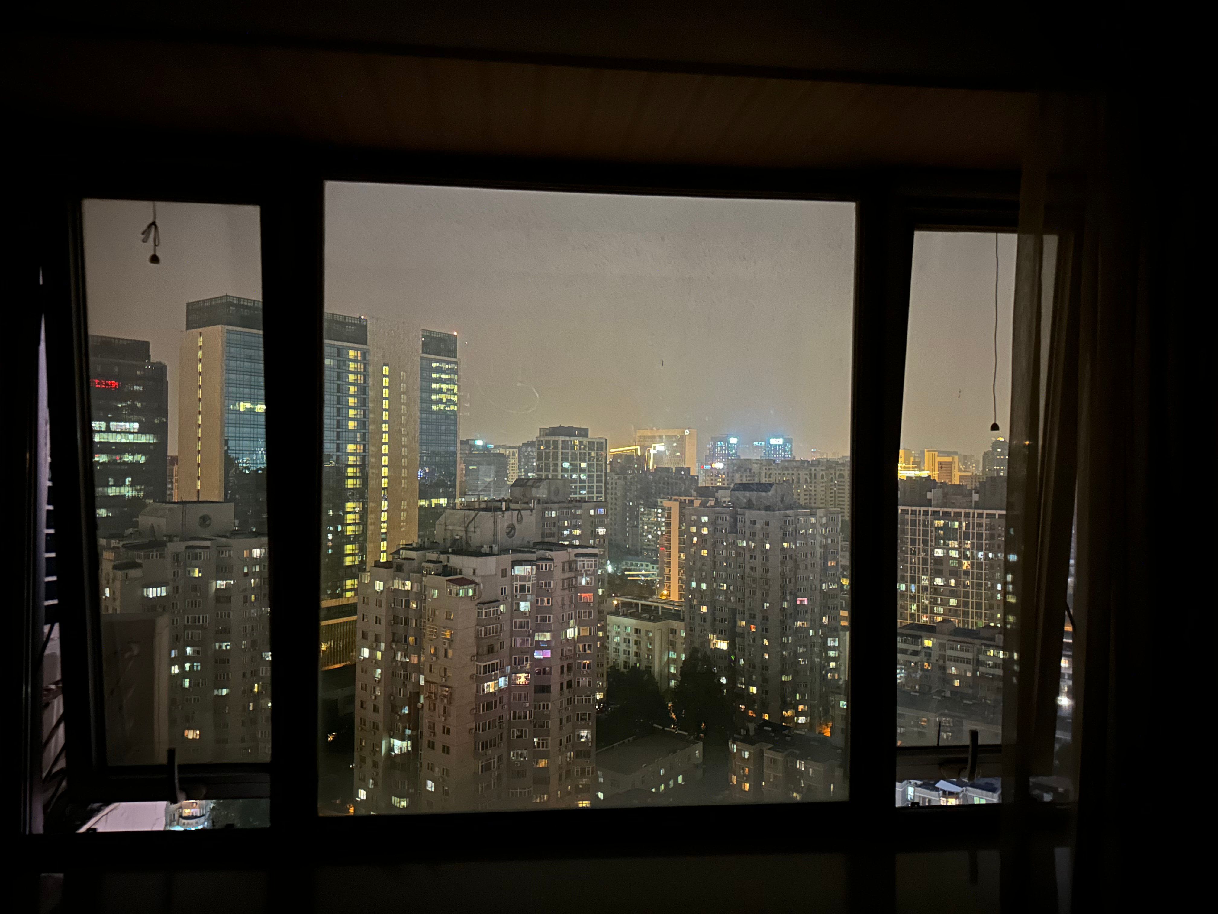 Der Blick aus dem Fenster eines Hotels in Beijing, der beleuchtete Hochhäuser der Stadt bei Nacht zeigt. Copyright: Yannic Han Biao Federer