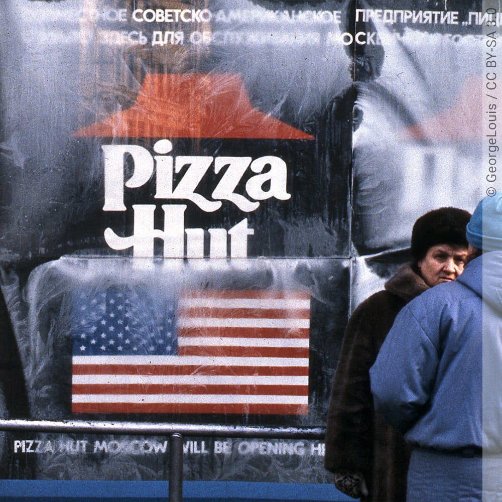 Ein Werbeplakat, abgebildet ist das Pizza Hut-Logo mit Schriftzug und die Flagge der Vereinigten Staaten, darunter steht "Pizza Hut Moscow", darüber ein Schriftzug in kyrillischer Schrift, zwei Personen in Winterkleidung stehen vor dem Plakat