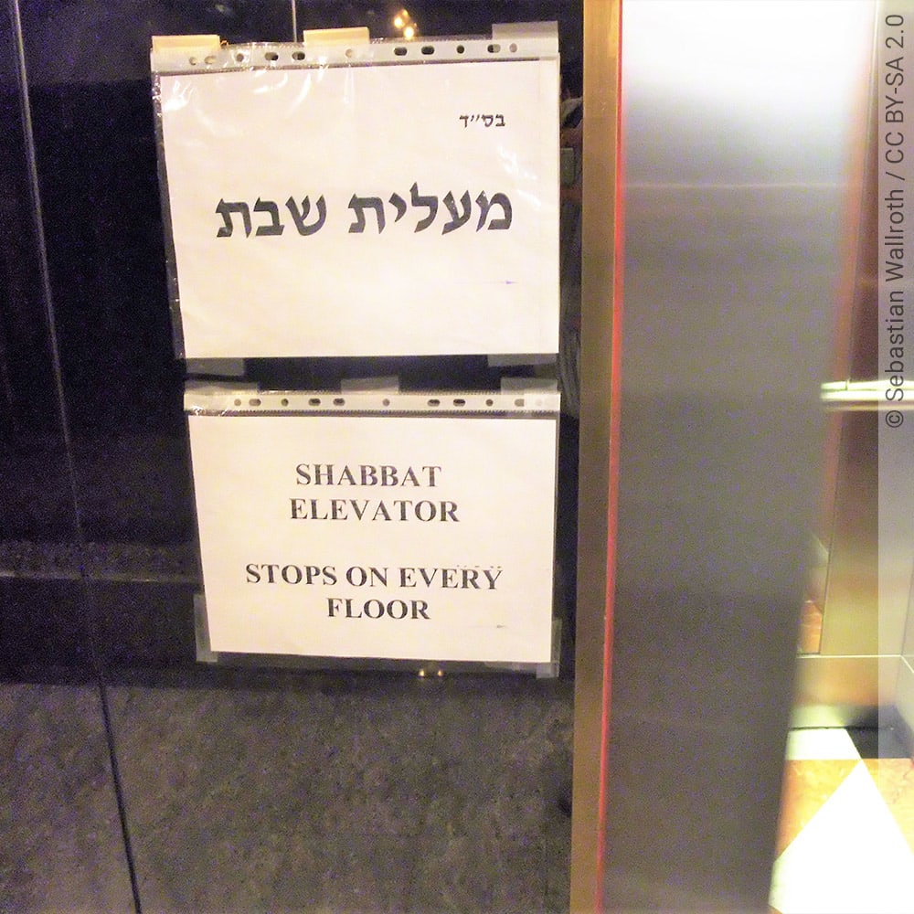 Zwei selbstgemachte Papierschilder an einem Shabbat-Aufzug, das obere Schild mit hebräischem Schriftzug, das untere Schild mit englischem Schriftzug "SHABBAT ELEVATOR STOPS ON EVERY FLOOR"