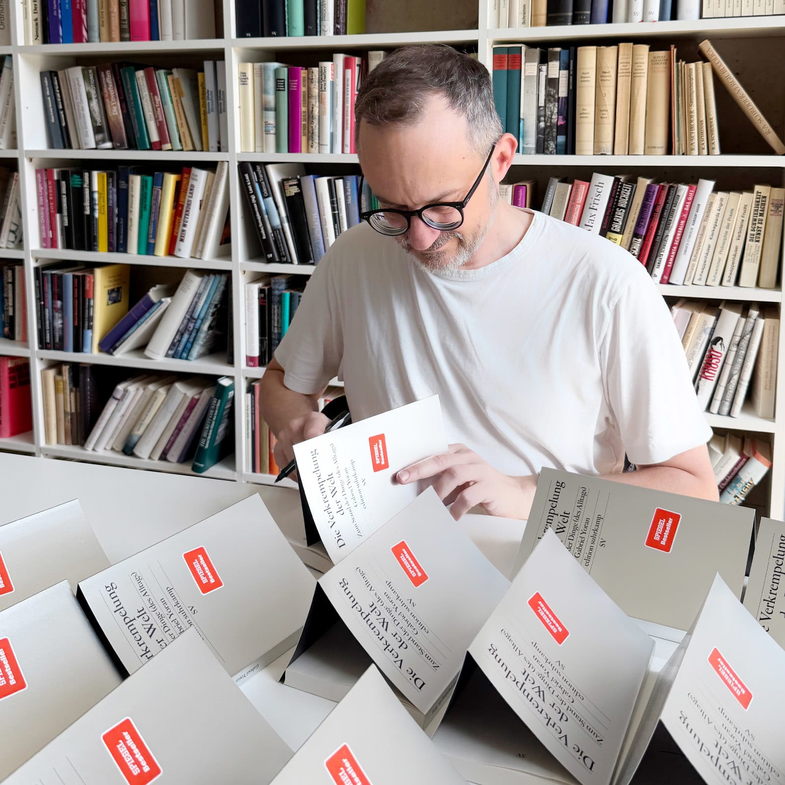 Gabriel Yoran sitzt vor einer Bücherwand und vor im liegen zahlreiche Bücher, die er signiert. 