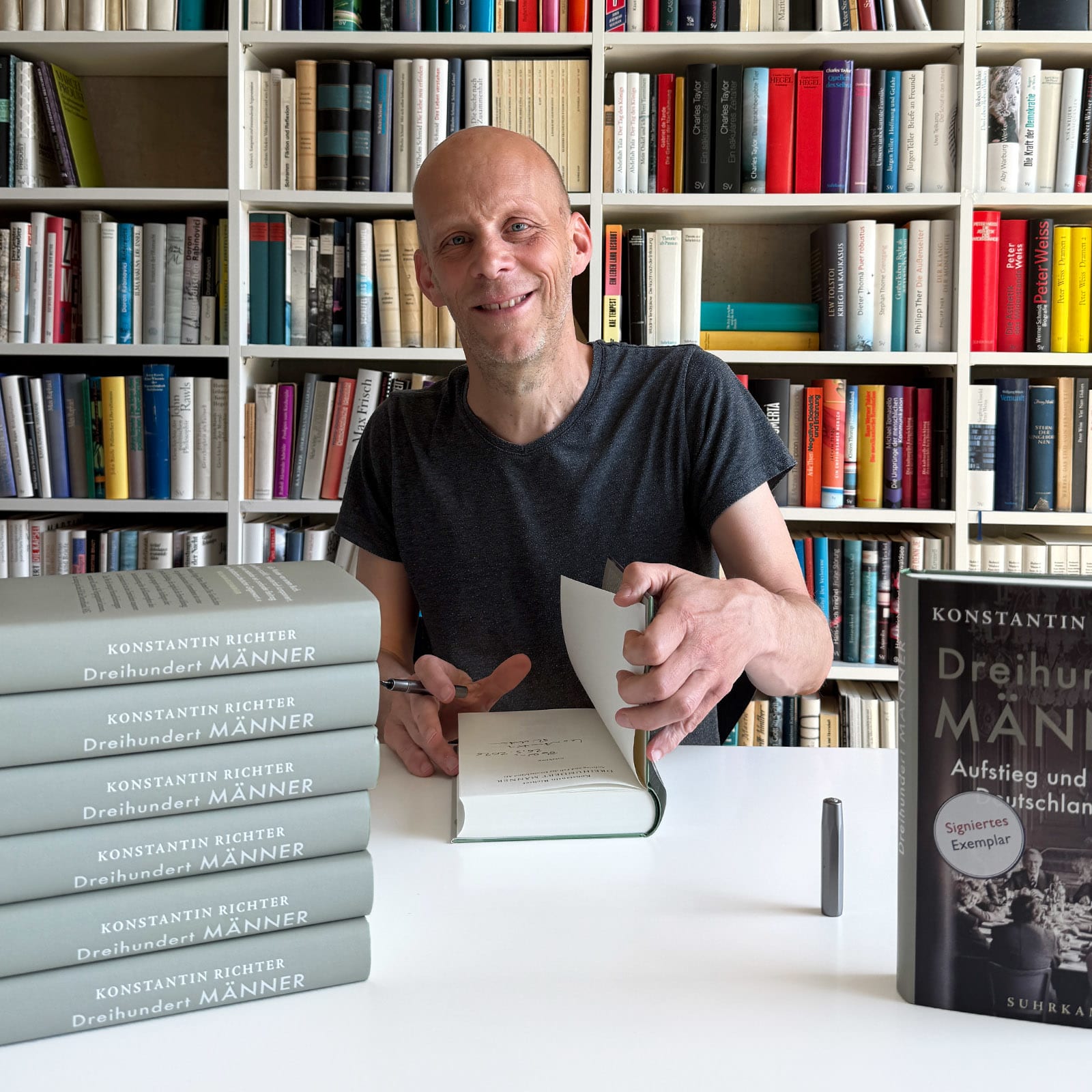 Konstantin Richter sitzt an einem Tisch und signiert sein Buch »Dreihundert Männer«, im Hintergrund ein Bücherregal.